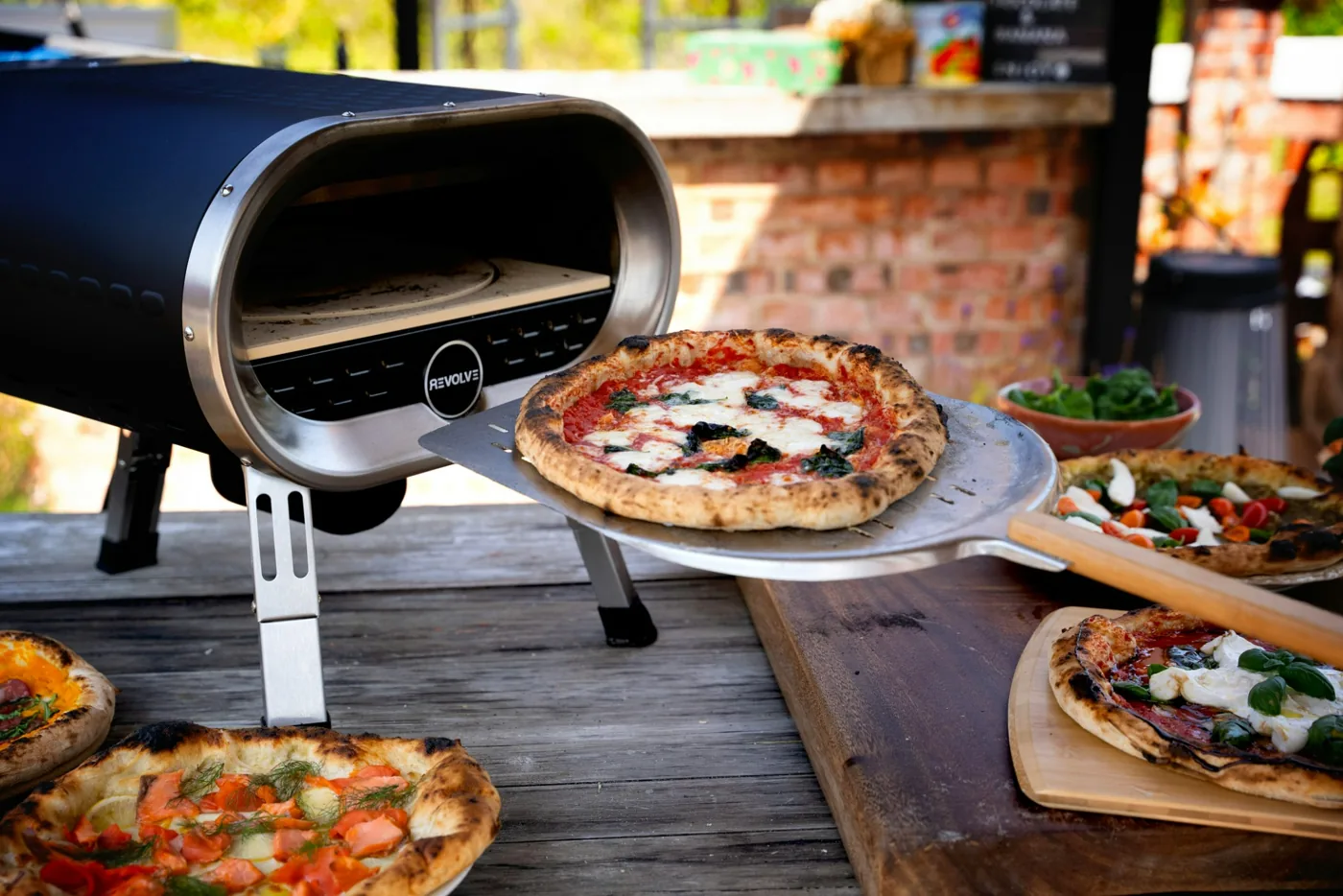 Cooked pizzas beside a portable outdoor pizza oven on a wooden table