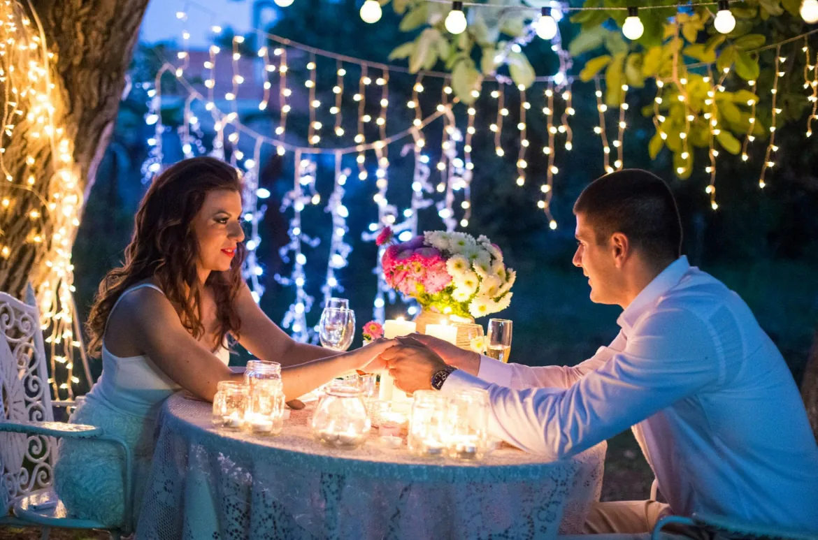 A couple holding hands across a candlelit table beneath strings of fairy lights