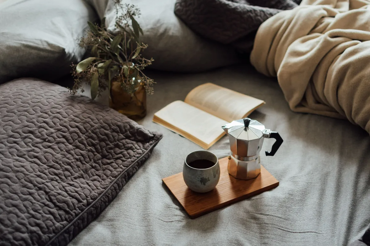 An open book, a coffee pot and a cup on a bed
