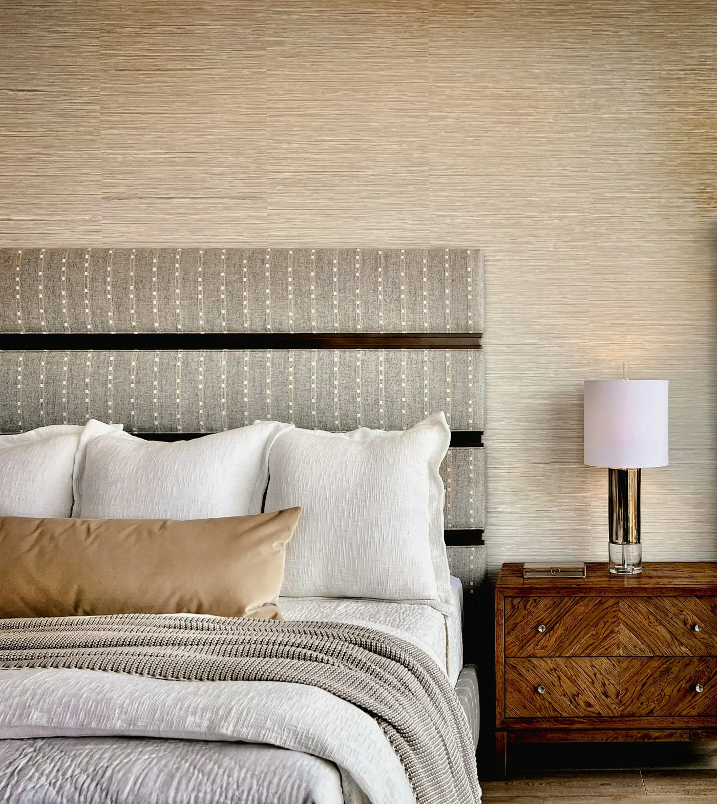 A made bed with white and grey bedding, cushions and a knitted throw beside a wooden bedside table