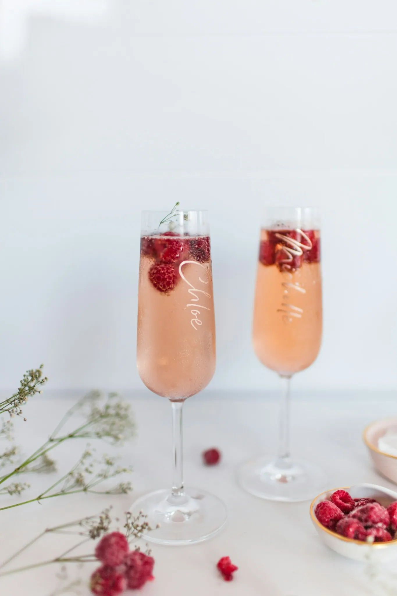 Two champagne flutes filled with a pink drink and raspberries, with pale script on the glass