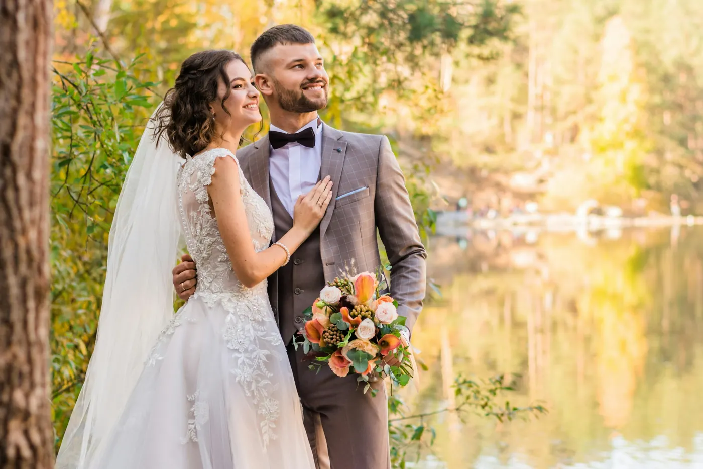 A bride and groom smiling together beside a lake, the bride holding a flower bouquet
